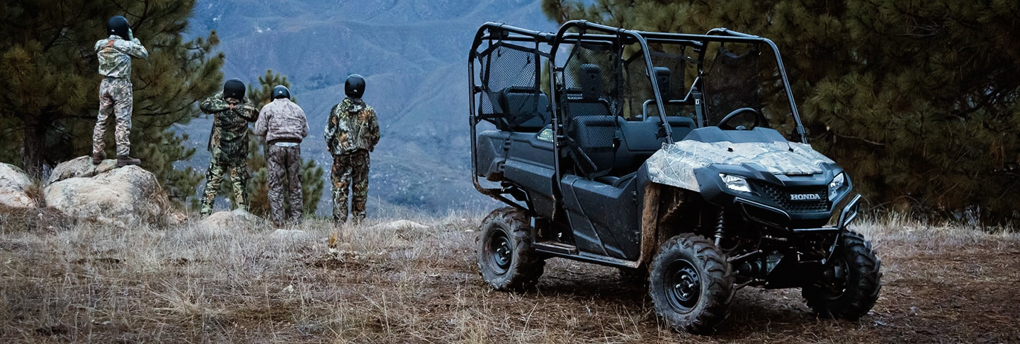 Pioneer 700-4 side-by-side parked in grassy wooded area with four ATV riders dressed in black helmets and camoflauge looking out over a hilly ridge in the background