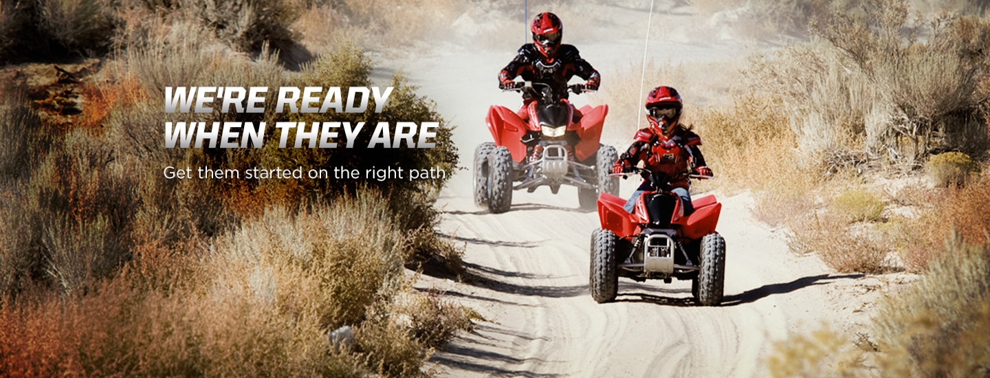 We're ready when they are. Get them started on the right path. Image of young child riding red ATV down dusty trail with adult riding behind in matching ATV